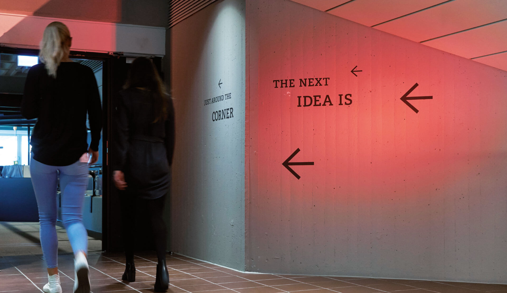 Siemens Coworking Space Beschriftungen mit Pfeilen und der typo THE NEXT IDEA IS an einer rot beleuchteten Beton Wand mit Personen daneben.