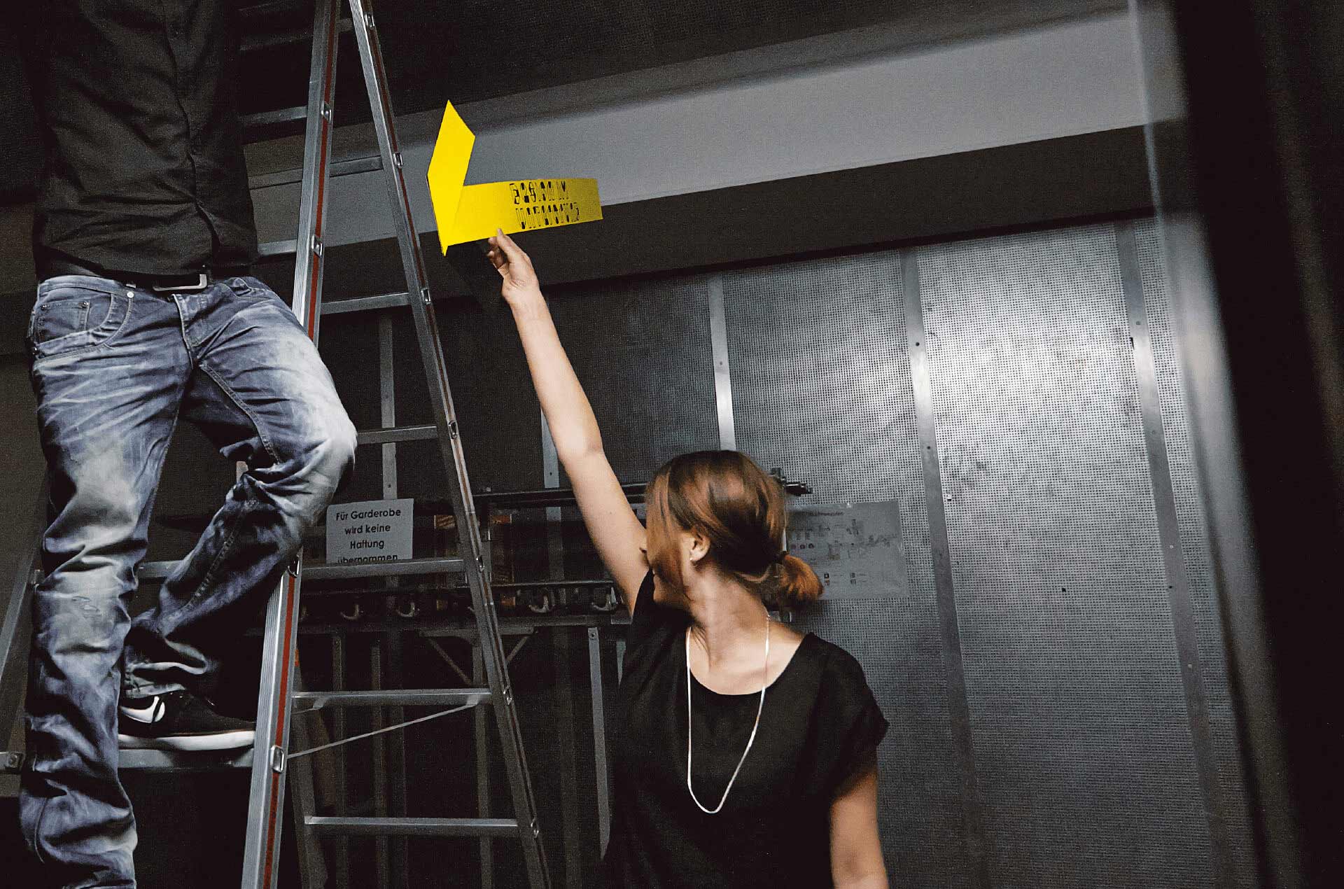 Frau beim Event Aufbau reicht Mann auf Leiter ein gelbes Papier mit Schriftzug