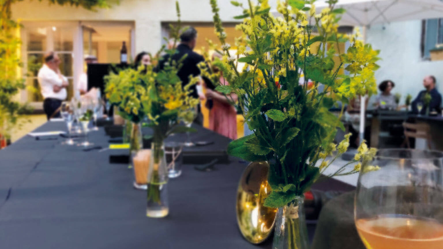 eine lange Tafel mit Getränken und Blumen im Hintergrund reden Menschen