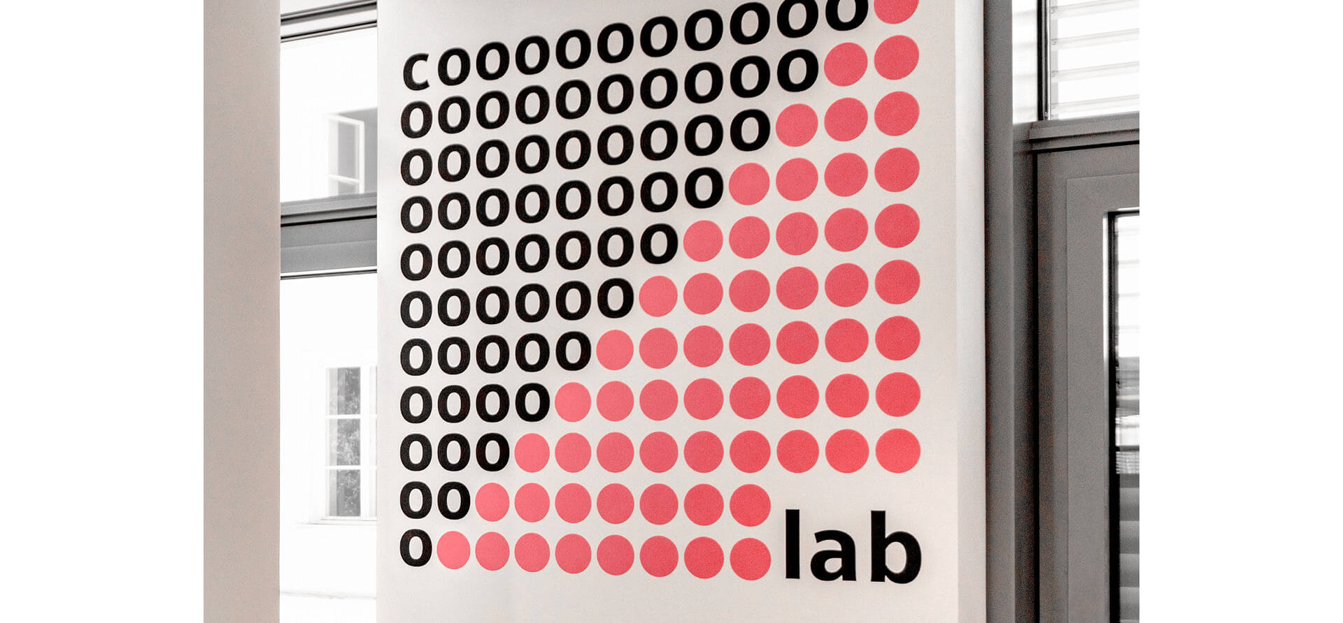 Raumgestaltungselement, auf dem das Wort co.lab dekorativ mit mehreren rosaroten Punkten und schwarzer Schrift auf weißem Hintergrund dargestellt wird. Im Hintergrund sieht man eine Fensterfront.