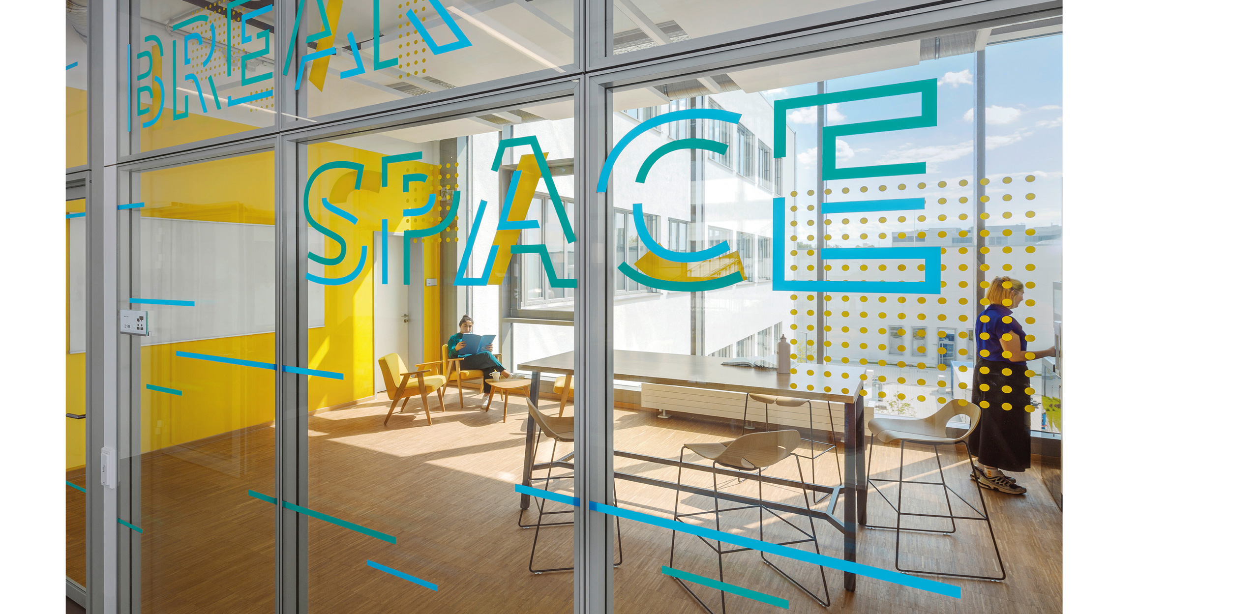 Break Space mit gelber Wand hinter Verglasung mit grünen und blauen Designelementen